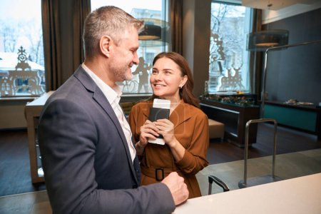 Photo for Adult smiling caucasian couple of woman with passport and man looking at each other at reception desk during check-in in modern hotel lobby. Concept of rest, vacation and travelling - Royalty Free Image