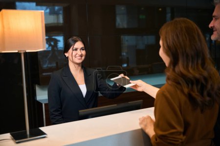 Photo for Smiling female european receptionist taking passport from adult woman near her cropped caucasian man at reception desk during check-in in hotel lobby. Concept of rest, vacation and travelling - Royalty Free Image