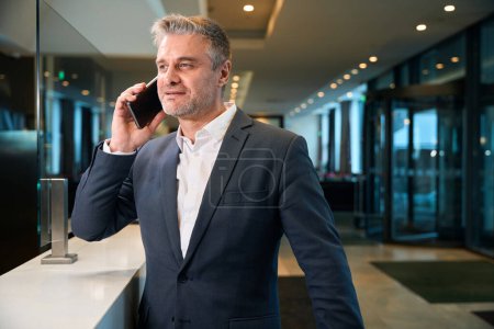 Foto de Hombre caucásico adulto concentrado hablando en el teléfono inteligente en recepción en el vestíbulo del hotel. Concepto de descanso, vacaciones y viajes - Imagen libre de derechos
