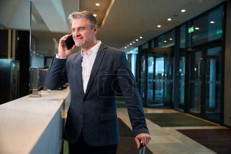 Photo for Focused adult caucasian man with suitcase talking on smartphone at reception desk in hotel lobby. Concept of rest, vacation and travelling - Royalty Free Image