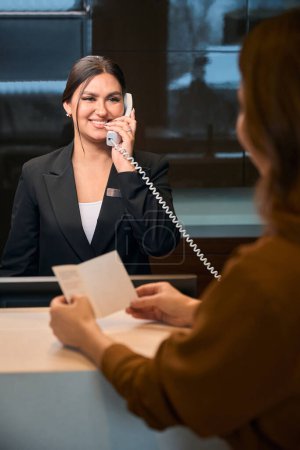 Foto de Mujer recortada con pasaporte y recepcionista europea sonriente hablando por teléfono mirándose durante el check-in en recepción en el vestíbulo del hotel. Descanso, vacaciones y viajes - Imagen libre de derechos