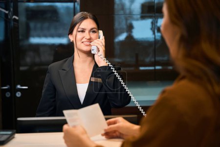 Téléchargez les photos : Femme partielle avec passeport et réceptionniste caucasienne souriante appelant au téléphone se regardant lors de l'enregistrement à la réception dans le hall de l'hôtel. Repos, vacances et voyages - en image libre de droit