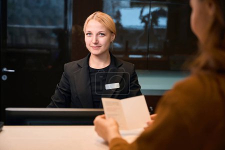 Téléchargez les photos : Femme partielle avec passeport près d'une réceptionniste européenne souriante regardant la caméra lors de l'enregistrement à la réception dans le hall de l'hôtel. Concept de repos, vacances et voyages - en image libre de droit