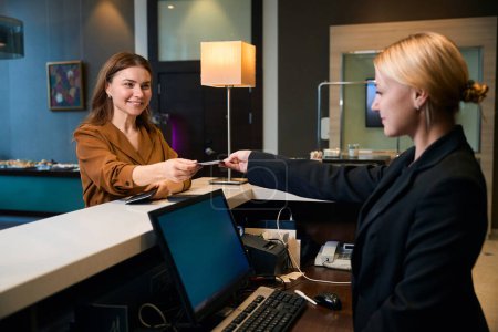 Photo for Smiling adult caucasian woman giving passport to young female european receptionist during check-in at reception desk in hotel lobby. Concept of rest, vacation and travelling - Royalty Free Image