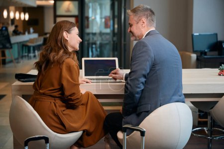 Photo for Back view of smiling adult caucasian couple sitting at wooden desk with laptop and looking at each other in hotel lobby. Concept of rest, vacation and travelling - Royalty Free Image