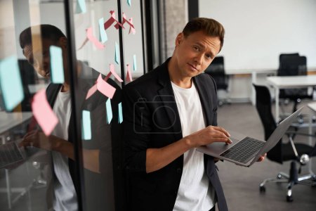 Photo for Self-confident handsome man looking at camera standing near the glass transparent wall with sticky notes and holding laptop, working on the move - Royalty Free Image