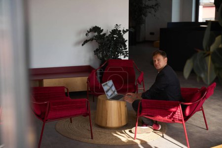 Téléchargez les photos : Homme travaillant ordinateur portable dans un coin confortable et confortable de la grande zone de travail de l'espace de coworking, assis dans un fauteuil rouge et regardant la caméra, paiement horaire et wi-fi gratuit - en image libre de droit
