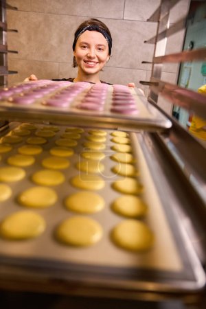 Téléchargez les photos : Joyeuse pâtissière mettant des moules à feuilles avec des coquilles de macaron sur un rack de rangement - en image libre de droit
