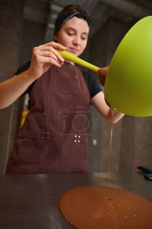 Téléchargez les photos : Confiseur concentré dans un tablier versant du chocolat fondu du bol sur le comptoir de la cuisine - en image libre de droit