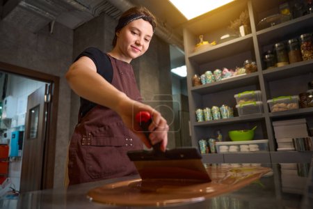 Téléchargez les photos : Jeune chocolatier souriant en tablier mélangeant chocolat fondu sur plan de travail avec spatule - en image libre de droit