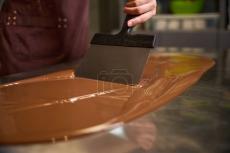 Photo for Cropped photo of confectioner in apron mixing melted chocolate on worktop with spatula - Royalty Free Image
