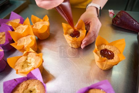 Foto de Foto recortada de panadero experimentado apretando mermelada de bolsa de plástico en el centro de cupcake - Imagen libre de derechos