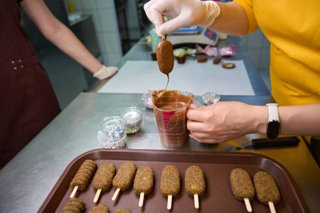 Photo for Cropped photo of confectioner dipping cakesicles into melted chocolate in presence of colleague - Royalty Free Image
