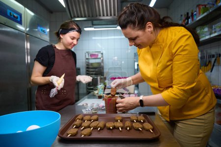 Photo for Smiling pastry chef dipping cake popsicles into melted chocolate in measuring cup in presence of her busy colleague - Royalty Free Image