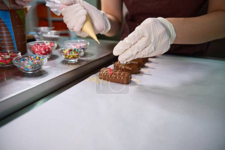 Photo for Cropped photo of confectioner in disposable gloves covering batch of cakesicles with sprinkles on kitchen countertop - Royalty Free Image