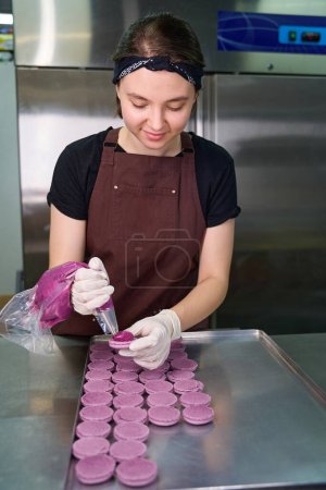 Téléchargez les photos : Pâtissier en gants jetables tuyauterie pourpre remplissage sur coquille de macaron sur plaque sur comptoir de cuisine - en image libre de droit