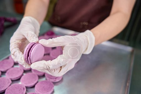 Photo for Cropped photo of pastry cook in disposable gloves assembling purple macaron shells together over baking tray on kitchen countertop - Royalty Free Image