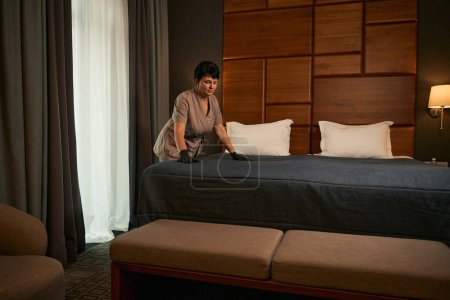 Focused maid in disposable gloves and uniform making bed with bedspread in suite