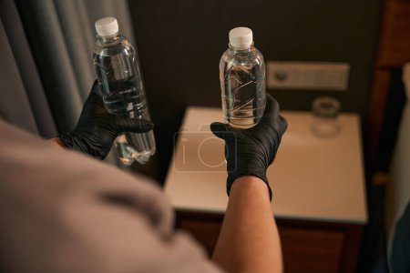 Foto de Foto recortada de la camarera en manos enguantadas colocando dos botellas de agua en la mesita de noche en suite - Imagen libre de derechos