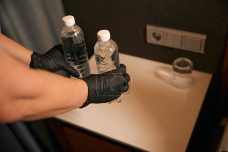 Foto de Primer plano de las manos femeninas en guantes desechables poniendo botellas de agua mineral en la mesita de noche en la habitación del hotel - Imagen libre de derechos
