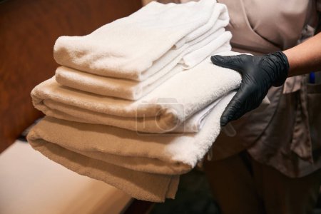 Téléchargez les photos : Photo recadrée de la femme de ménage dans des gants en nitrile noir jetables tenant pile de serviettes de bain blanches propres - en image libre de droit