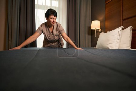 Téléchargez les photos : Femme de chambre en uniforme lissant la surface du couvre-lit avec les mains pendant la fabrication du lit dans la suite d'invités - en image libre de droit