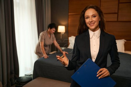Photo for Smiling room service supervisor with pen and clipboard in hands posing for camera while chambermaid making bed on background - Royalty Free Image