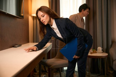 Téléchargez les photos : Superviseur de ménage inspectant la table pour la propreté en présence de la bonne occupée dans la suite - en image libre de droit
