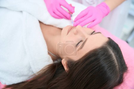 Téléchargez les photos : Patient couché sur le canapé, cosmétologue tenant la seringue et faisant une injection dans les lèvres. Augmentation des lèvres - en image libre de droit