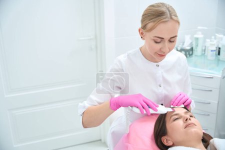 Photo for Young lady lying on couch, beautician holding device and making ultrasonic face cleaning to client - Royalty Free Image