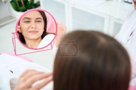 Téléchargez les photos : Patient souriant tenant un miroir rose et regardant la peau, attendant la procédure - en image libre de droit