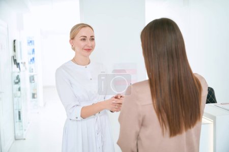 Téléchargez les photos : Cosmétologue professionnel en manteau blanc debout près de la réception et parlant avec la femme après la procédure - en image libre de droit