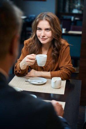 Téléchargez les photos : Femme heureuse tenant une tasse dans la main alors qu'elle était assise à la table dans un café - en image libre de droit