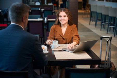 Photo for Cheerful woman sitting at the table with coffee and laptop on it while listening to a man opposite her - Royalty Free Image