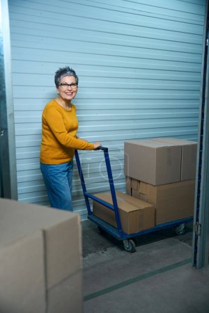 Photo for Adult woman in glasses transports things in a cardboard box on a blue cart - Royalty Free Image