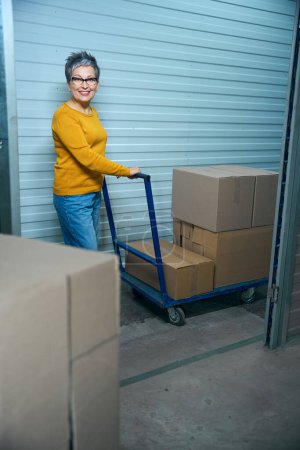 Photo for Adult woman holds a cart for transporting things on which there are cardboard boxes. - Royalty Free Image