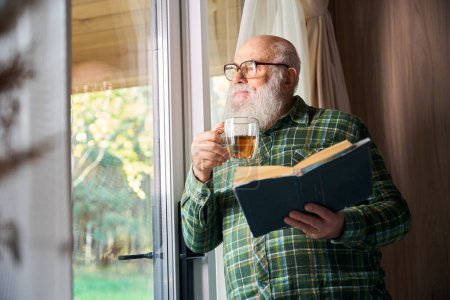 Téléchargez les photos : Un homme âgé prend une tasse de thé près de la fenêtre, il a un livre à la main - en image libre de droit