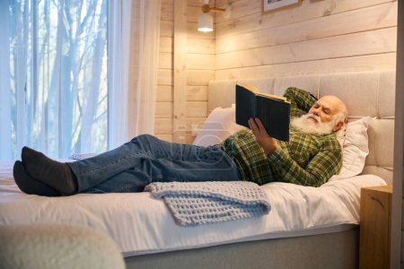 Téléchargez les photos : Vieil homme allongé avec un livre sur un lit confortable, la chambre a un intérieur minimaliste - en image libre de droit