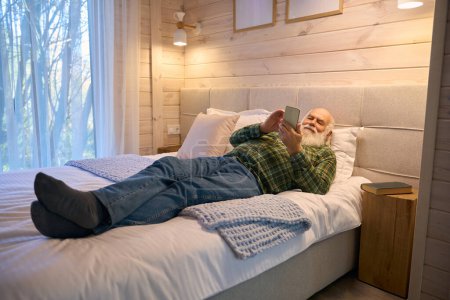 Téléchargez les photos : Le vieil homme était fatigué et allongé pour se reposer sur un lit confortable, il avait un téléphone portable dans ses mains - en image libre de droit
