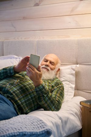 Foto de Pensionista está cansado y se acuesta a descansar en una cama acogedora, tiene un teléfono móvil en sus manos - Imagen libre de derechos