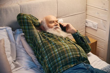 Foto de Viejo se acuesta a descansar en una cama acogedora, se comunica en su teléfono móvil - Imagen libre de derechos