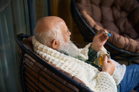 Téléchargez les photos : Vieil homme avec une boisson fraîche thé chaud sur la véranda, il a un thermomètre dans ses mains - en image libre de droit