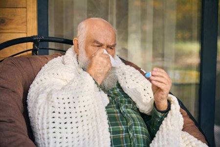 Foto de El viejo enfermo está envuelto en una mullida manta en la terraza, tiene un termómetro en sus manos - Imagen libre de derechos