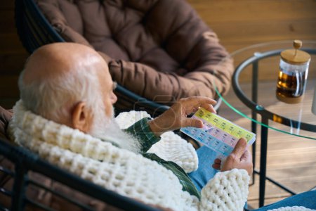 Foto de El pensionista está envuelto en una mullida manta en la terraza, sosteniendo una caja de pastillas en sus manos - Imagen libre de derechos