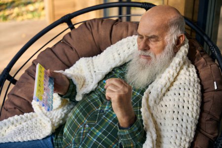 Photo for Pensioner is wrapped in a fluffy blanket, he has a pill box and a pill in his hands - Royalty Free Image