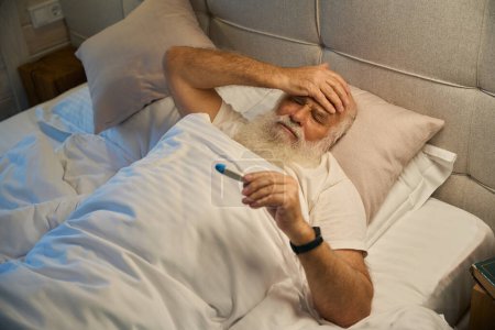Foto de El viejo sufre de frío y dolor de cabeza, tiene un termómetro en las manos - Imagen libre de derechos