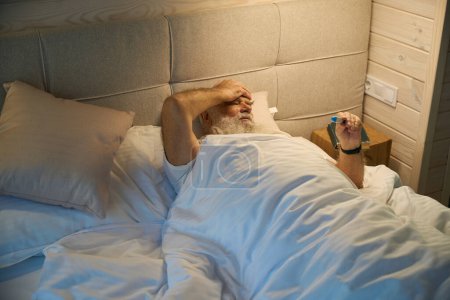 Foto de El viejo mide la temperatura corporal con un termómetro, tiene un resfriado y dolor de cabeza - Imagen libre de derechos