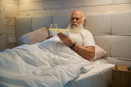 Foto de El viejo sostiene el pillbox y un vaso de agua en sus manos, está sentado en una cama grande - Imagen libre de derechos