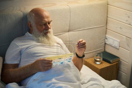 Foto de Anciano sostiene una caja de tabletas y una píldora en sus manos, que se encuentra en una cama grande - Imagen libre de derechos