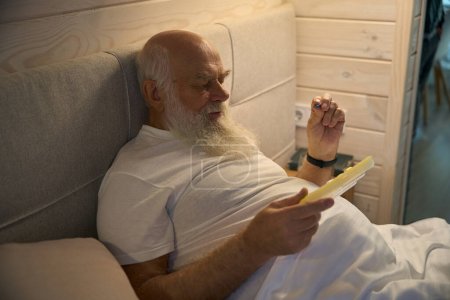 Photo for Old man holds a tablet box and a pill in his hands, he is sitting on a large bed - Royalty Free Image